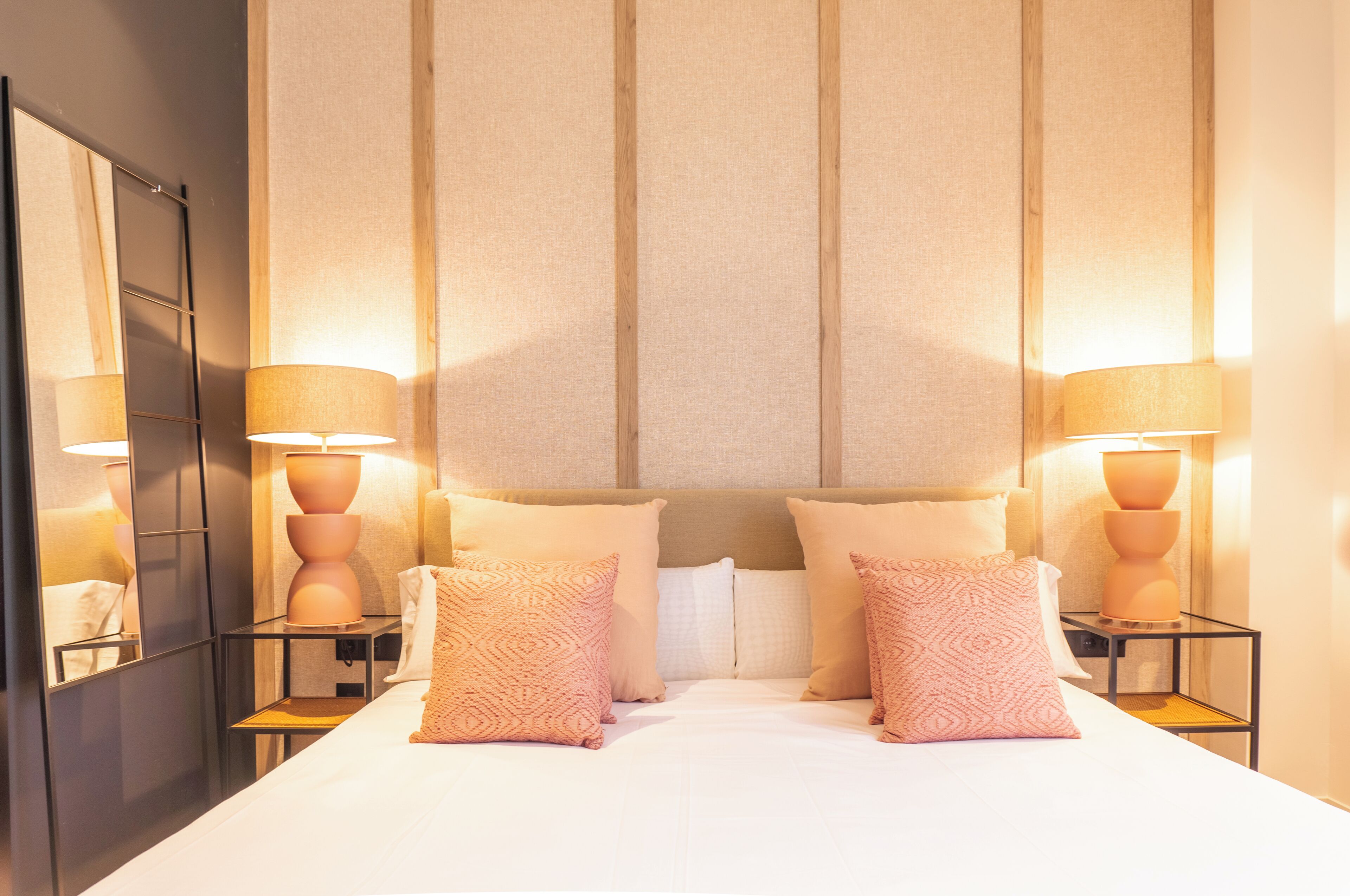 Una habitación de hotel con una cama grande, dos mesitas de noche con lámparas, un espejo y un cabecero con líneas verticales.