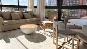 A modern living room with a sofa, round coffee table, wicker chairs, and a dining table. Large windows offer a view of the cityscape. - Valencia Luxury - Calma Beach Apartments (Valencia)