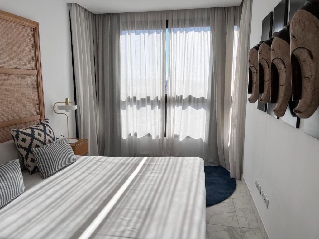 Un dormitorio con una cama grande, una mesita de noche, una lámpara y vistas al exterior a través de cortinas translúcidas.