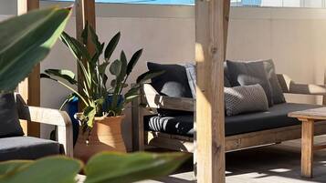 A wooden outdoor seating area with a cushioned bench and a table, surrounded by potted plants.