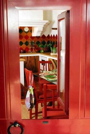 A red-framed doorway leading to a kitchen with a tiled backsplash, a clock, and a dining area with a table and chairs.
