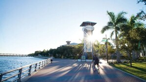 A waterfront promenade with a tall tower, palm trees, and a bridge in the background.