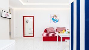 Ein modernes Hotelzimmer mit einer weißen und roten Sofagarnitur, einem Esstisch mit Frühstücksangeboten, einem großen Spiegel und einem gerahmten Kunstwerk an der Wand.