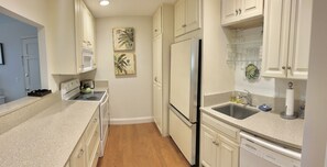 A compact kitchen with white cabinetry, a stainless steel refrigerator, a dishwasher, and a microwave. There are two wall-mounted shelves with glassware and a small plant.