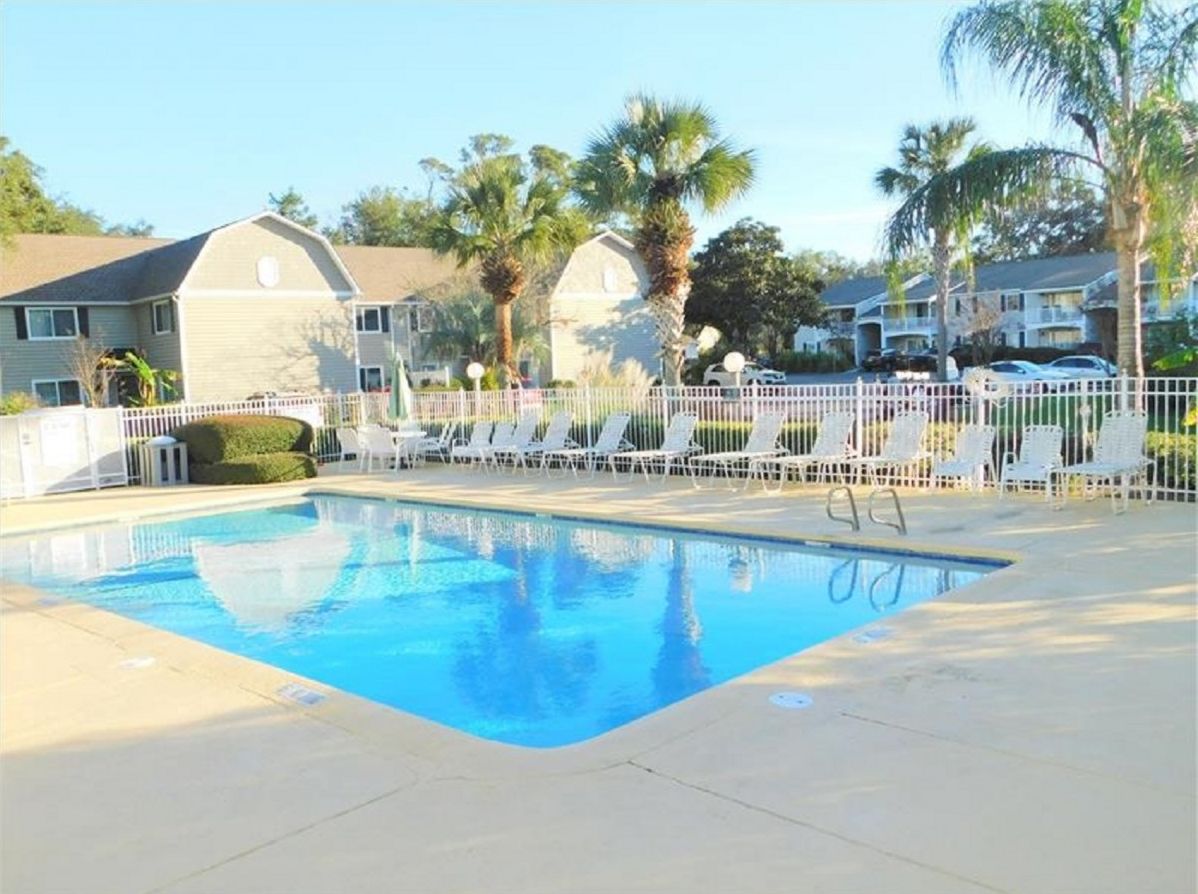 A swimming pool with lounge chairs and a fence, surrounded by palm trees and residential buildings.