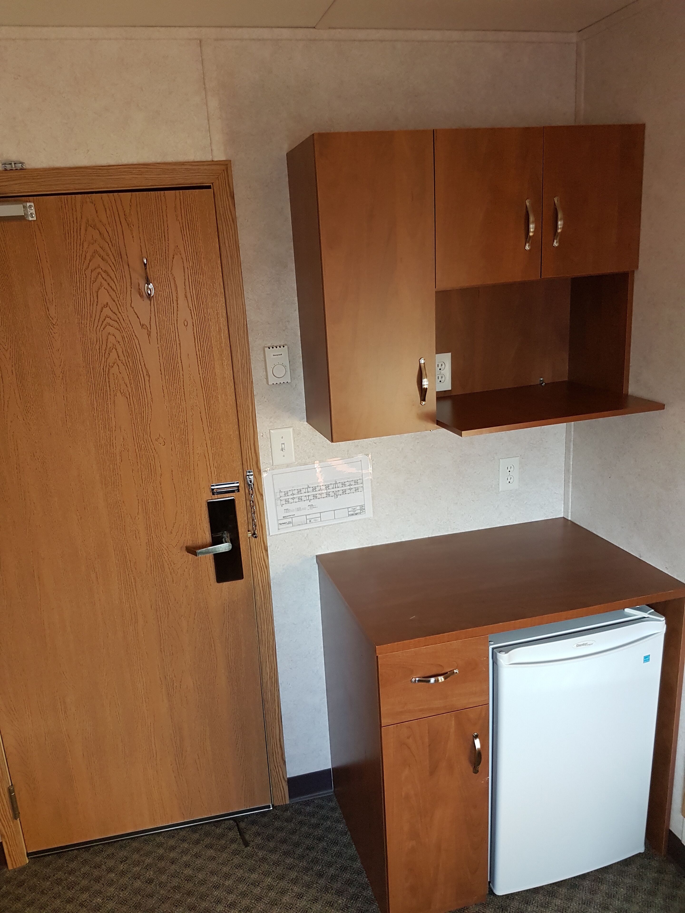 A compact kitchenette with a wooden door, a small refrigerator, and a wooden cabinet above the counter.