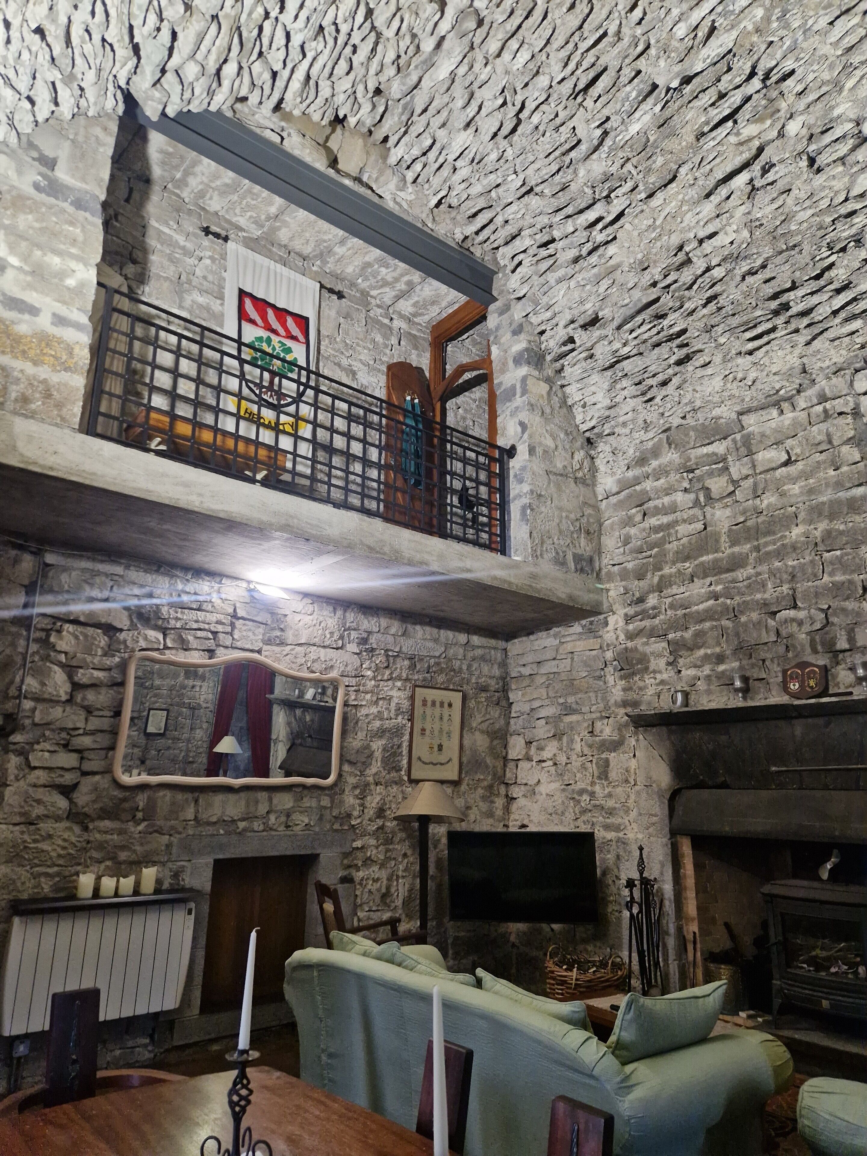 A rustic room with exposed stone walls, a wooden ceiling, a balcony with a metal railing, a fireplace, and a sofa.