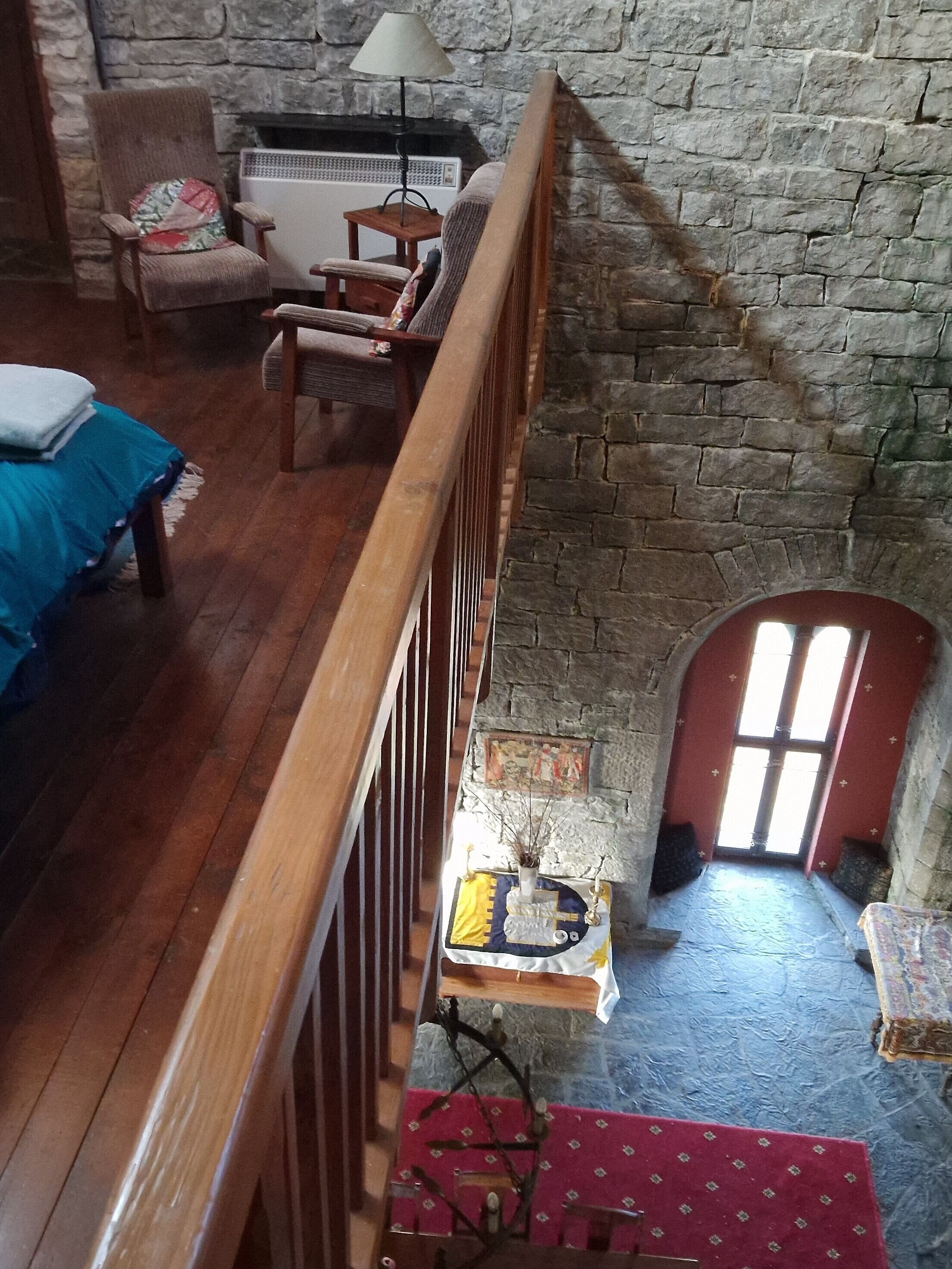 Une pièce avec un plancher en bois, un mur en pierre, une rambarde en bois, un lit recouvert d’une courtepointe bleue, une chaise avec un coussin, une petite table avec une lampe et une fenêtre donnant sur l’extérieur.