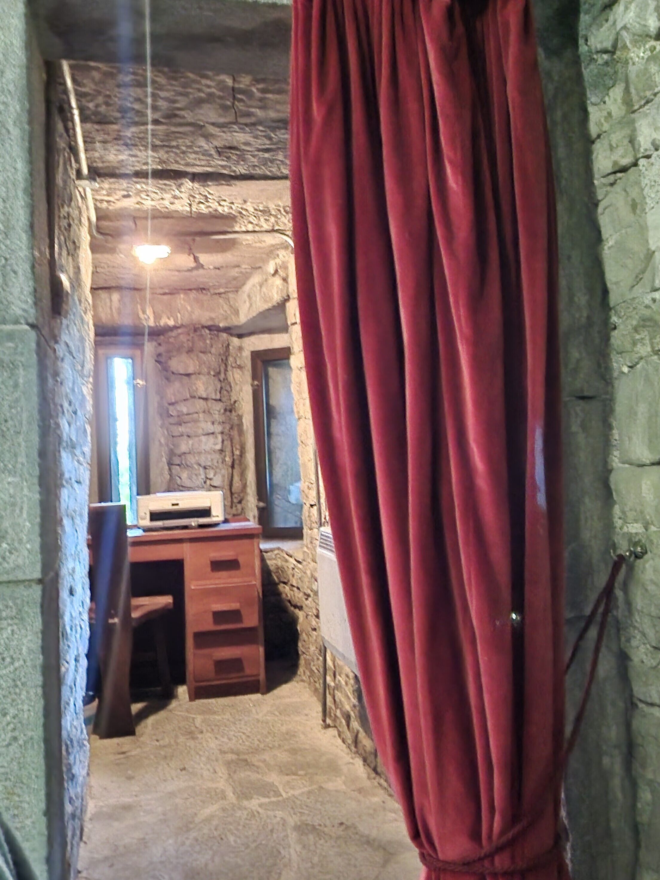 A narrow stone-walled corridor with a wooden desk and drawers, a red curtain, and a hanging light fixture.