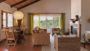 A cozy living room with a fireplace, a dining area with a table and chairs, and large windows offering a view of the outdoors. - Partridge House (Coruche)