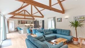 Een ruime woonkamer met een grote blauwe bank, houten balken in het plafond, een keuken op de achtergrond en een potplant.
