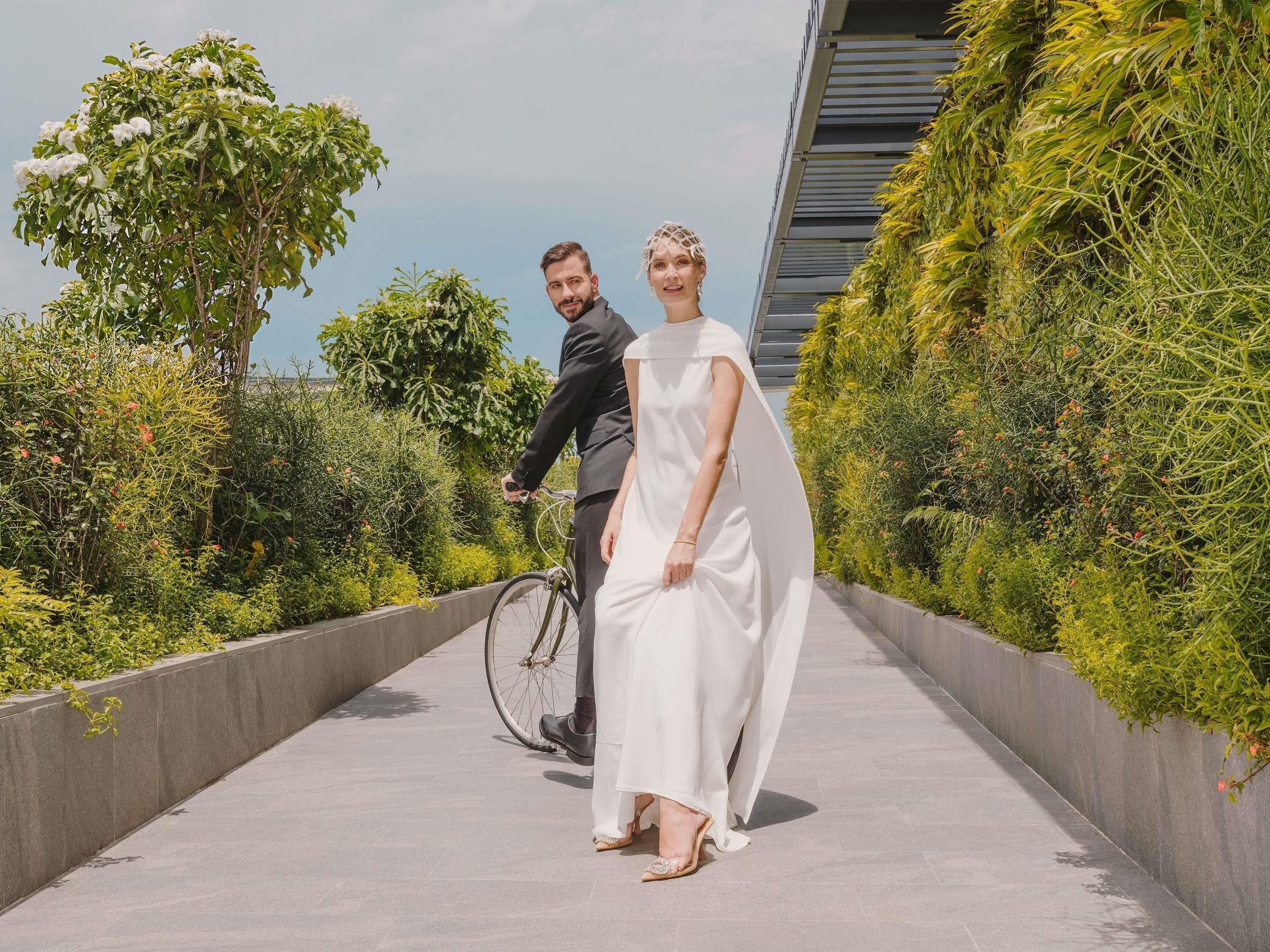 A couple on a paved pathway with greenery on either side, a bicycle, and a modern building in the background.
