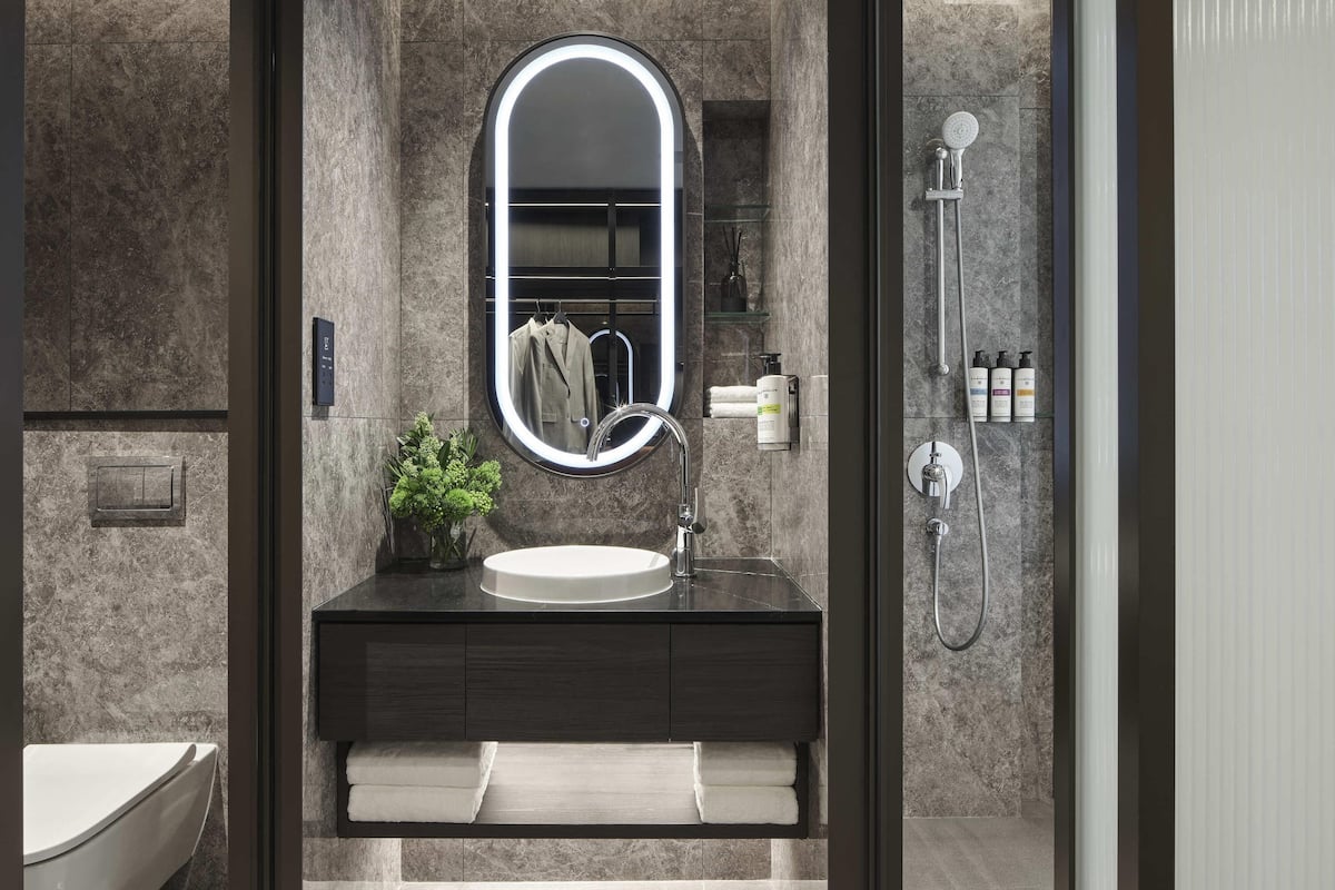 A modern bathroom with a marble wall, a round mirror with a light, a sink, a towel rack, and a shower.