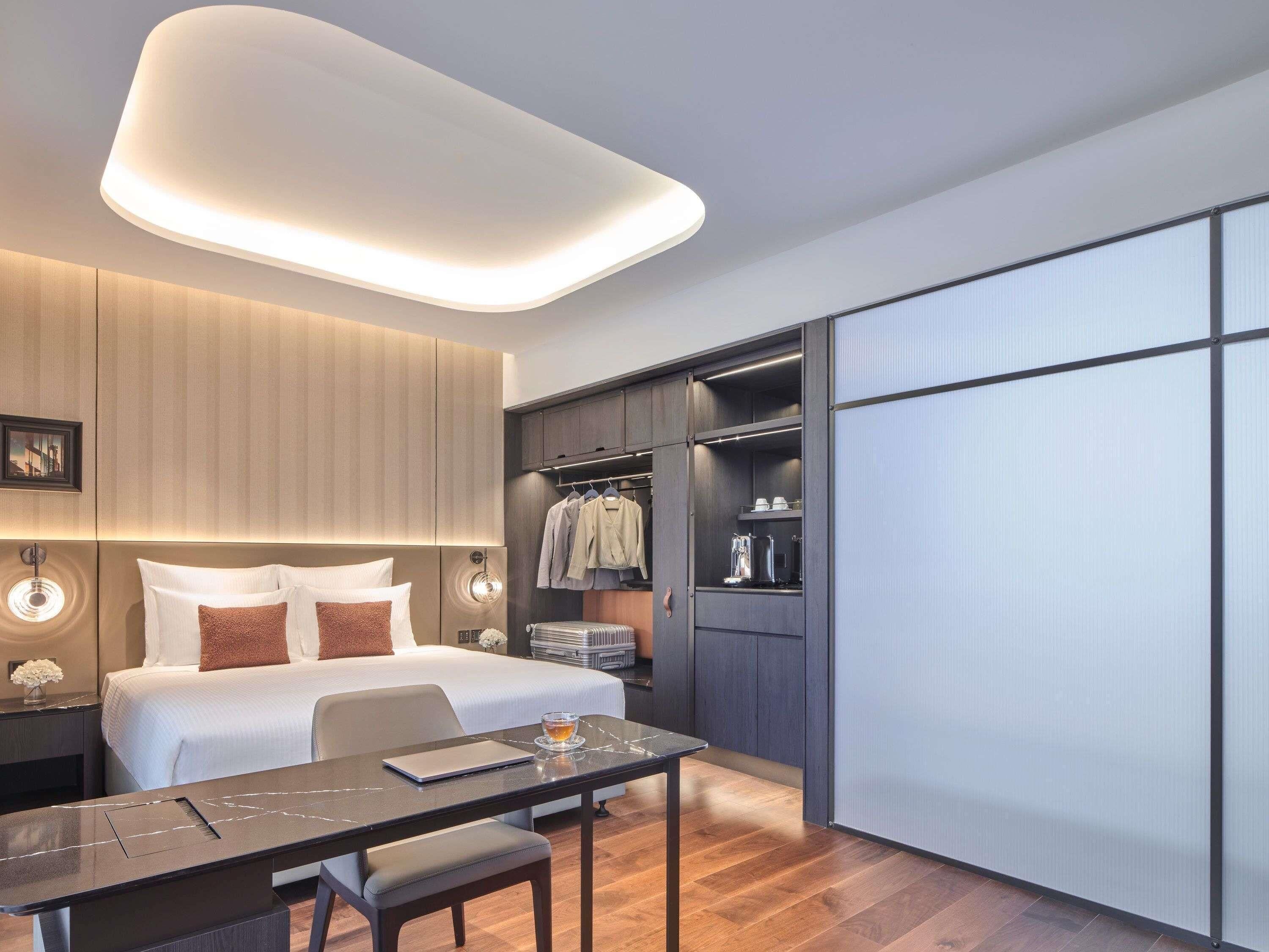 Ein modernes Hotelzimmer mit Bett, Nachttisch, Schreibtisch, Stuhl und begehbarem Kleiderschrank.