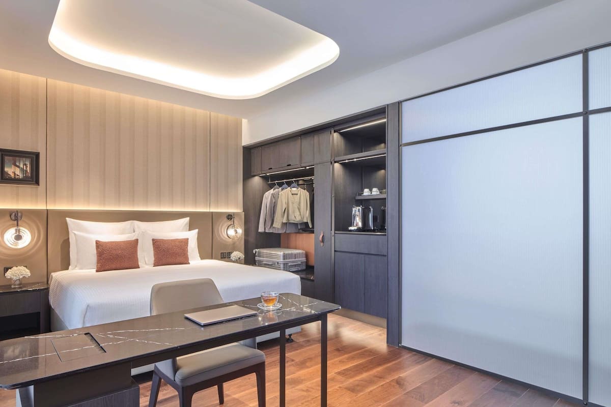 A modern hotel room with a bed, bedside table, desk, chair, and a walk-in closet.