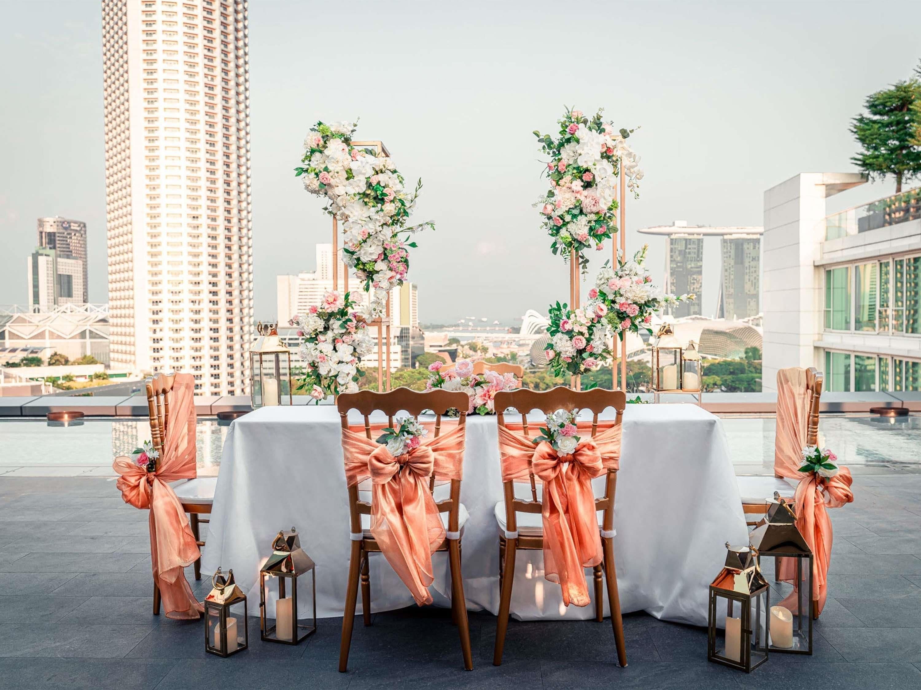 Un área de comedor en la azotea con una mesa dispuesta para cuatro personas, rodeada de sillas con cintas anaranjadas y arreglos florales.