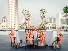 A rooftop dining area with a table set for four, surrounded by chairs with orange sashes, and floral arrangements.