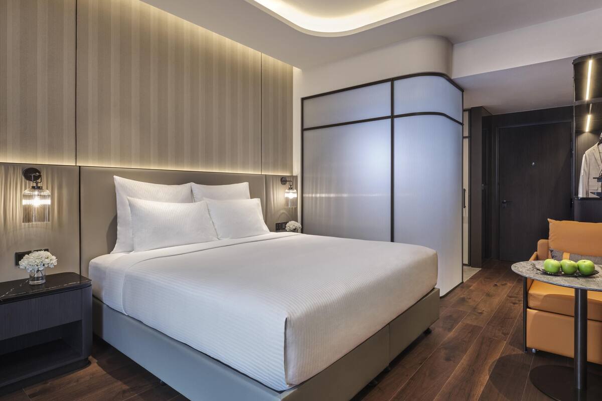 Una habitación de hotel moderna con una cama grande, una mesita de noche, un rincón de estar con una mesa y un armario.