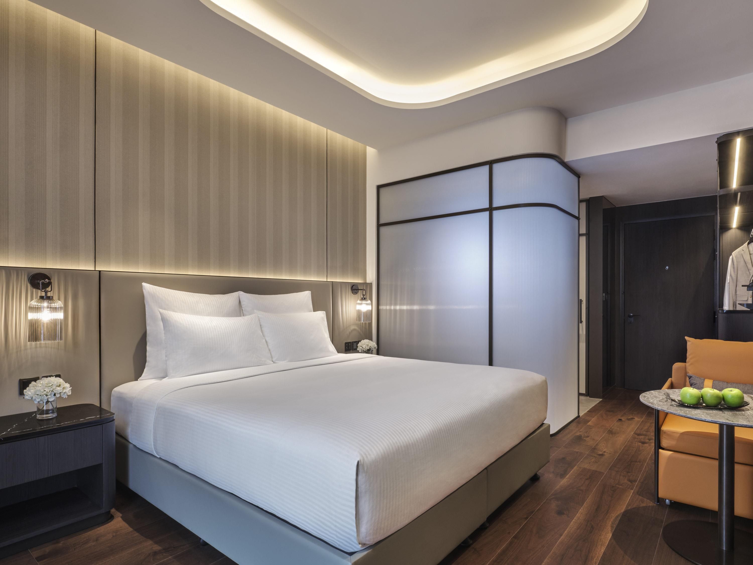A modern hotel room with a large bed, a nightstand, a seating area with a table, and a wardrobe.
