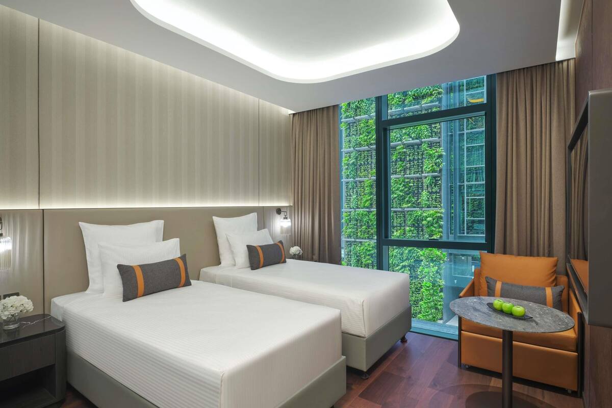 Una habitación de hotel moderna con dos camas, una mesita pequeña y vistas a zonas verdes a través de amplios ventanales.