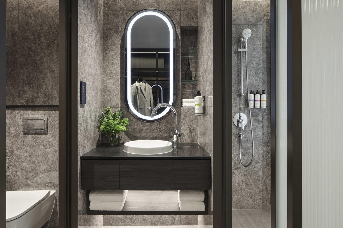 Un baño moderno con pared de mármol, un espejo redondo con luz, un lavamanos, un perchero y una ducha.