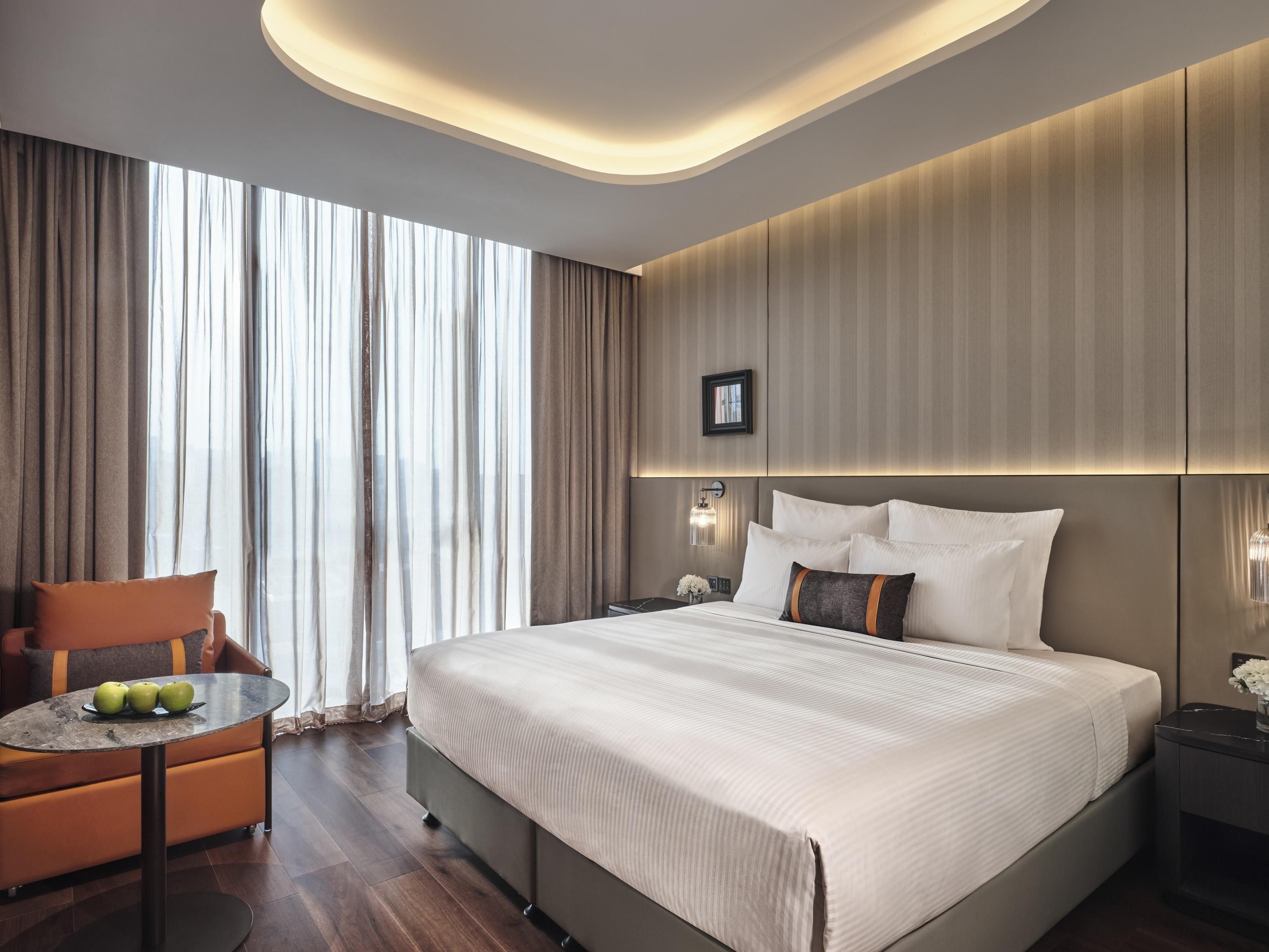 A modern hotel room with a large bed, a round table with apples, a chair, and a view through sheer curtains.