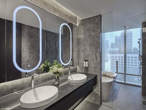 A modern bathroom with two sinks, a large mirror with illuminated edges, a vase with flowers, and a view of a cityscape through a glass door. - Pullman Singapore Hill Street (Singapore)