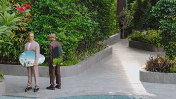 Deux personnes se tiennent près d’une piscine, entourées d’une végétation luxuriante, et on peut voir le reflet d’un paysage urbain dans un objet circulaire.