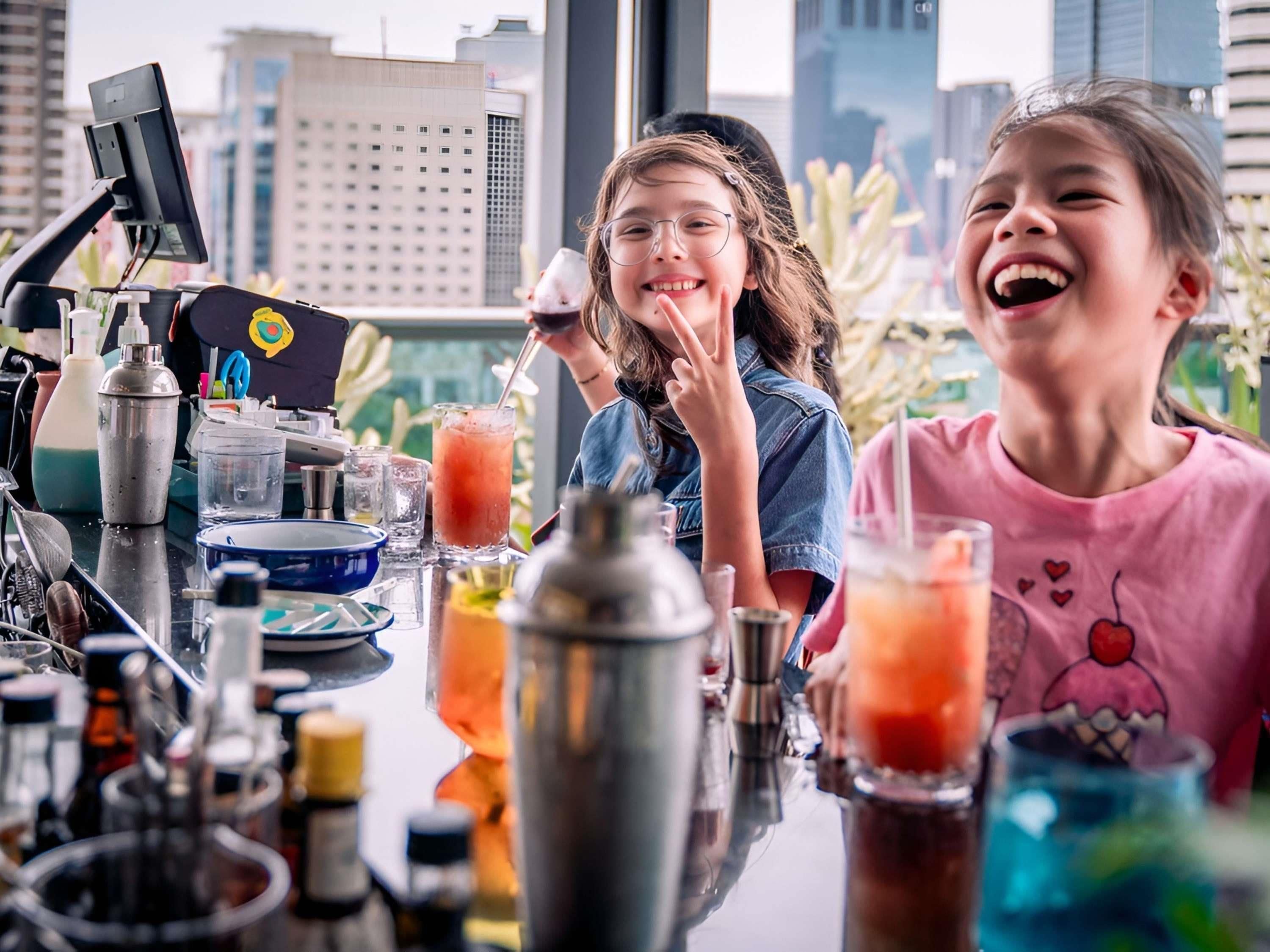 Een bar met verschillende cocktailshakers en glazen, een computer monitor en een stadsgezicht door grote ramen.