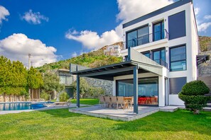 A modern two-story house with a covered patio, a swimming pool, and a view of a hillside.
