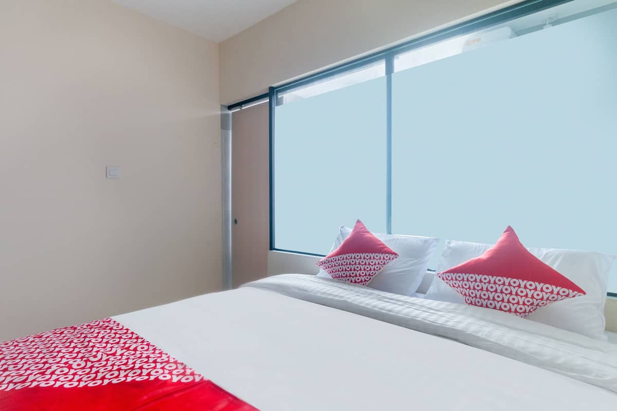 A hotel room with a large window, a bed with white bedding and red accents, and a red and white patterned throw blanket.