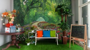 A cozy room with a mural of a forest, a white bench with colorful cushions, a small table with a vase of flowers, and a chalkboard menu. - OYO 3216 Ellegan Residence (South Tangerang)