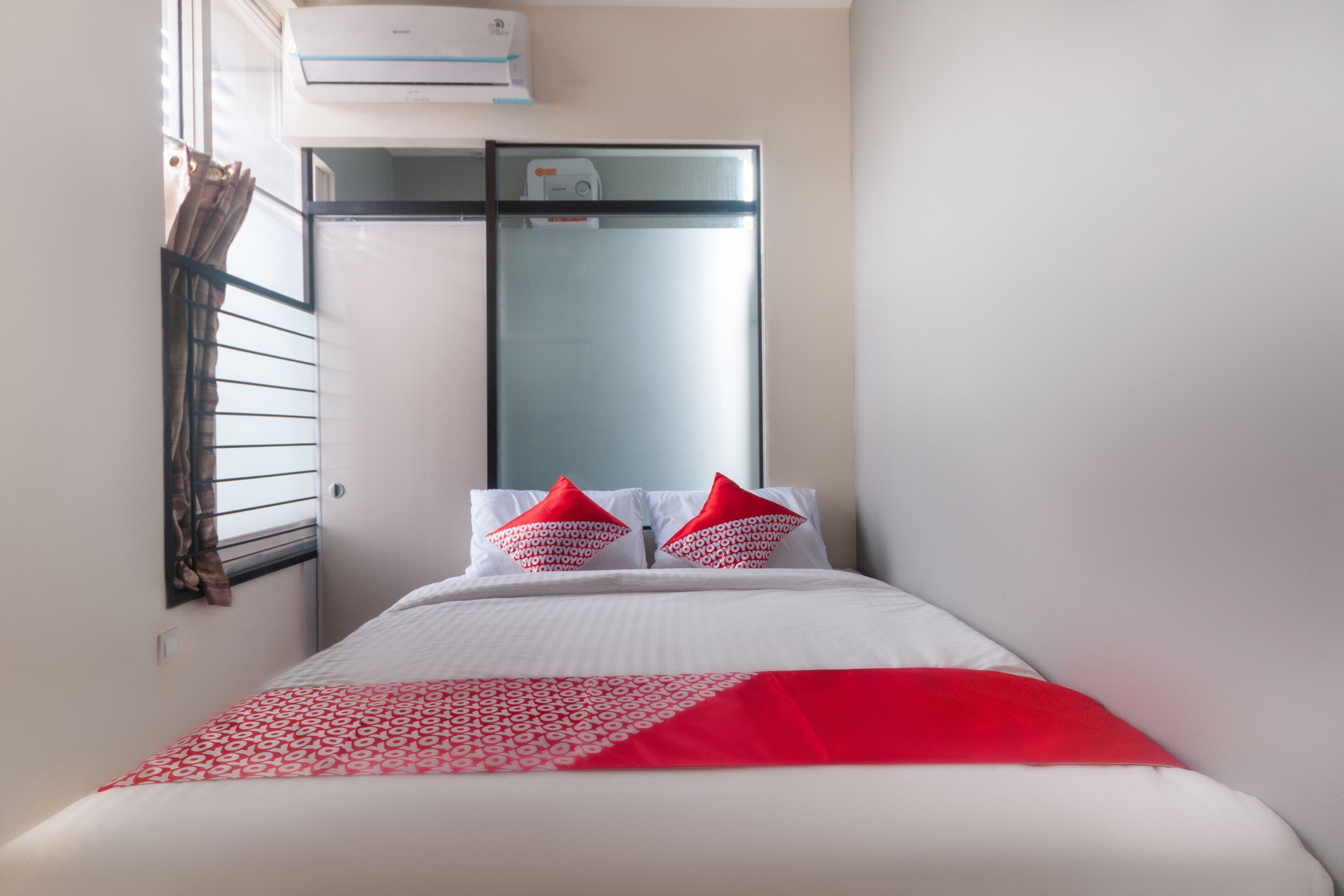 A hotel room with a bed, two red pillows, a white bedspread, and a window with blinds.