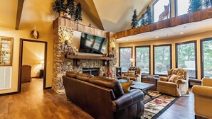 A spacious living room with a stone fireplace, leather sofas, and large windows offering views of the outdoors. - Little River Lodge sleeps 12 (Fort Payne)