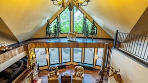 A spacious loft with a high ceiling, a fireplace, seating area, and a large window with a view of trees. - Little River Lodge sleeps 12 (Fort Payne)