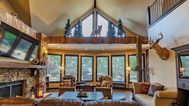 A spacious living room with a stone fireplace, leather sofas, and a large window with a view of trees. - Little River Lodge sleeps 12 (Fort Payne)