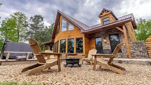 A log cabin with a stone foundation, large windows, and a patio with Adirondack chairs. - Little River Lodge sleeps 12 (Fort Payne)