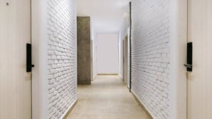 Un couloir aux murs en briques blanches et au sol carrelé, qui mène à une porte au fond.