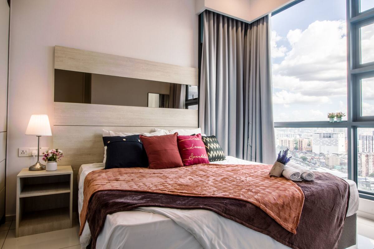Une chambre d’hôtel moderne avec un grand lit, une table de chevet avec une lampe et une vue sur la ville grâce à de grandes fenêtres.