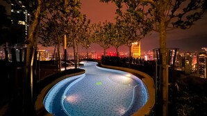 Une piscine nocturne offrant une vue sur le paysage urbain, entourée d’arbres et éclairée par un système d’éclairage artificiel.