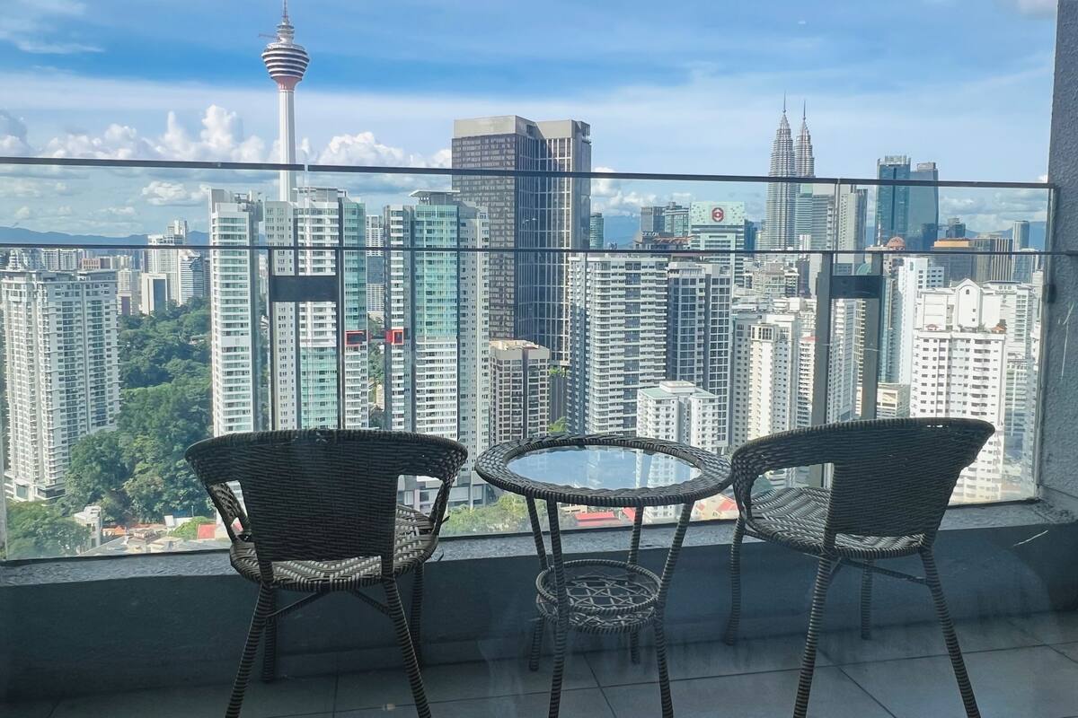 Un balcon agrémenté de chaises en osier et d’une petite table, d’où l’on peut admirer un panorama urbain avec ses gratte-ciel et son imposante tour.