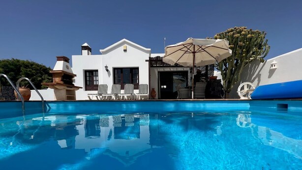 Ein klares, blaues Schwimmbecken mit einem weißen Gebäude im Hintergrund, eine Terrasse mit gestreiften Stühlen und einem großen Sonnenschirm.