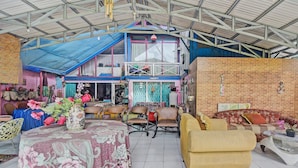 A spacious indoor area with a high ceiling, a blue roof, and a brick wall. There are several chairs and a sofa, a table with a floral tablecloth, and a vase with flowers. - SPOT ON 92278 Hotel Vorel (Tondano)