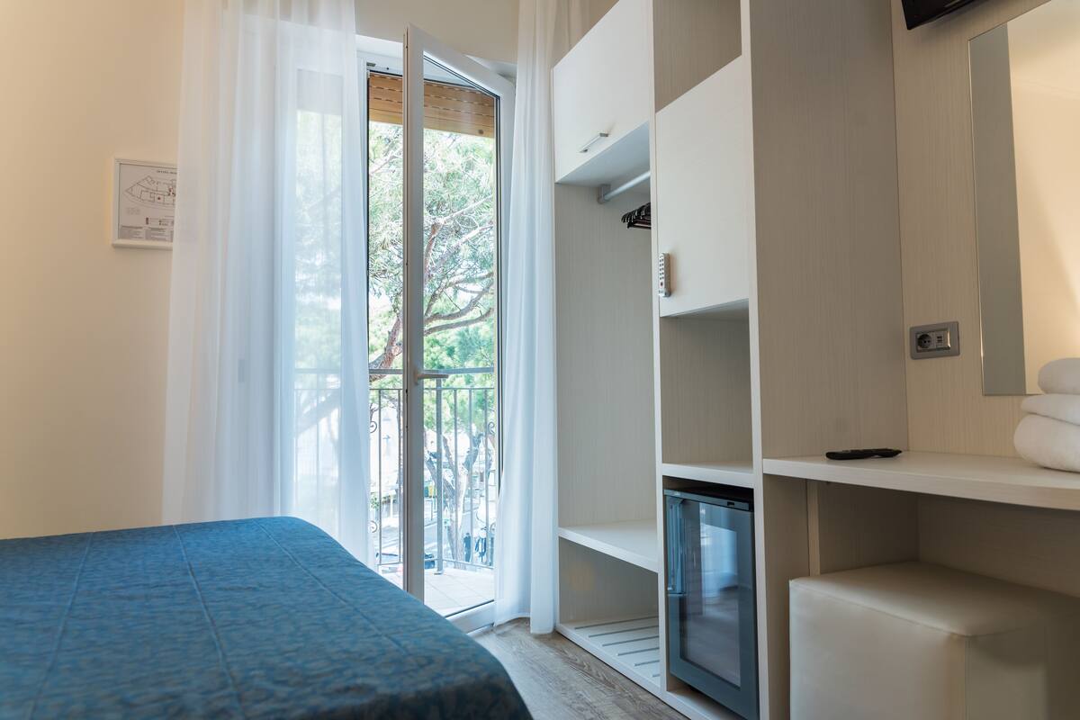 A bedroom with a bed, a built-in TV, a small refrigerator, and a balcony with a view of trees and a street.