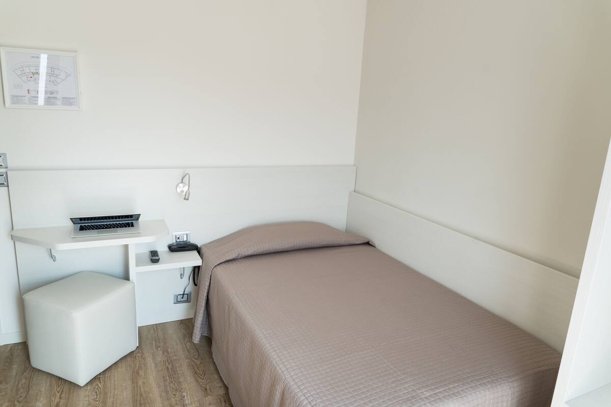 A small, neatly organized bedroom with a bed, a desk with a laptop, a chair, and a small table.