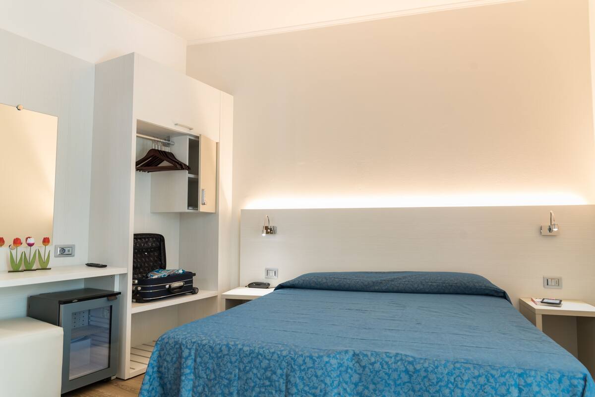 A hotel room with a bed, a desk, a wardrobe, and a small refrigerator.