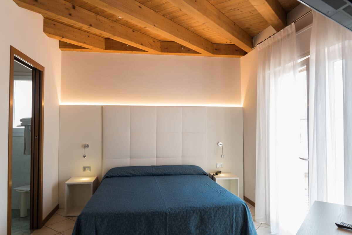 A bedroom with a wooden ceiling, a bed with a blue bedspread, bedside lamps, and a window with curtains.