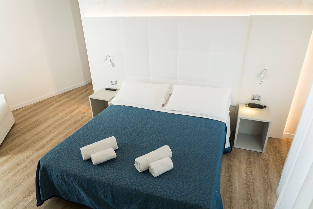 A neatly made bed with a blue bedspread and two rolled towels, a bedside table with a lamp, and a wooden floor.