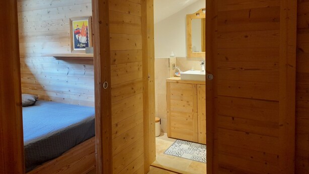 A wooden cabin interior with a bed, a bathroom with a sink and mirror, and a small shelf with a framed picture.