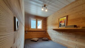 A small, cozy room with wooden walls, a bed with two pillows, a window, and a shelf with a picture.